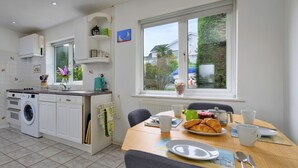 Una cocina con lavadora, una mesa de comedor con cosas para el desayuno y un ventanal con vistas a zonas verdes.