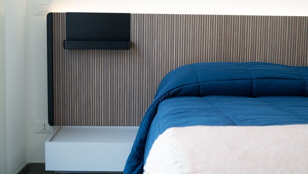 Un letto con coperta blu, un comodino bianco e una TV a parete.
