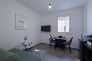A small, modern room with a green sofa, a small table with a vase of flowers, a dining table with chairs, a flat-screen TV, and a window with a view of the outside. - GuestHost - Cozy Apartment Pigneto Metro C (Roma)