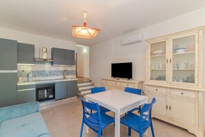 A modern kitchen with grey cabinets, a white dining table with blue chairs, a flat-screen TV, and a cabinet with glass doors. - Apartment 'Villino San Teodoro' with Private Terrace, Garden and Air Conditioning (San Teodoro)