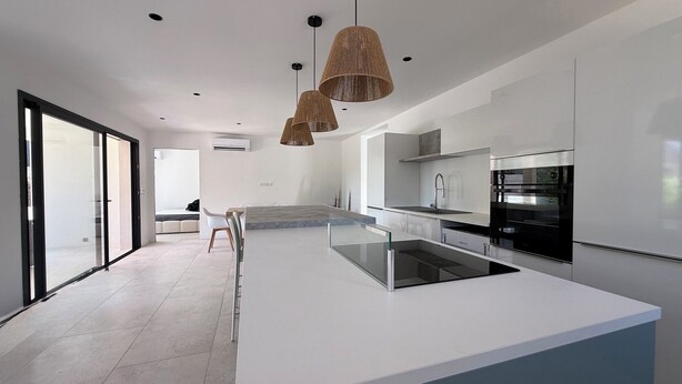 A modern kitchen with a central island, a dining area with a glass table, and a large sliding glass door leading to a balcony. - Magnificent luxury detached villa T4 private pool (Santa-Lucia-di-Moriani)