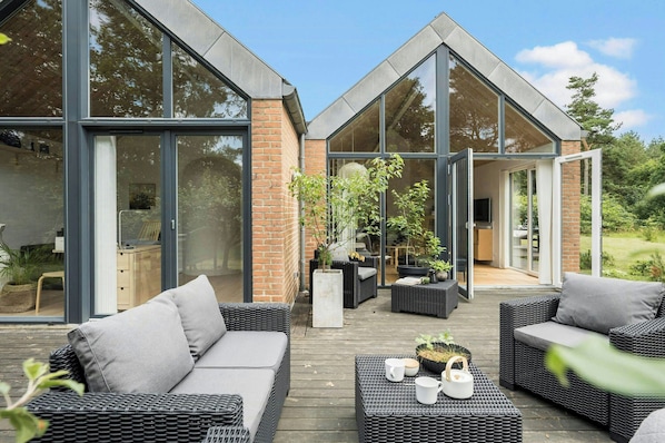 A modern outdoor living space with wicker furniture, a wooden deck, and glass doors leading to a garden. - Holiday home on a large natural lot by Hjarbæk (Skals)