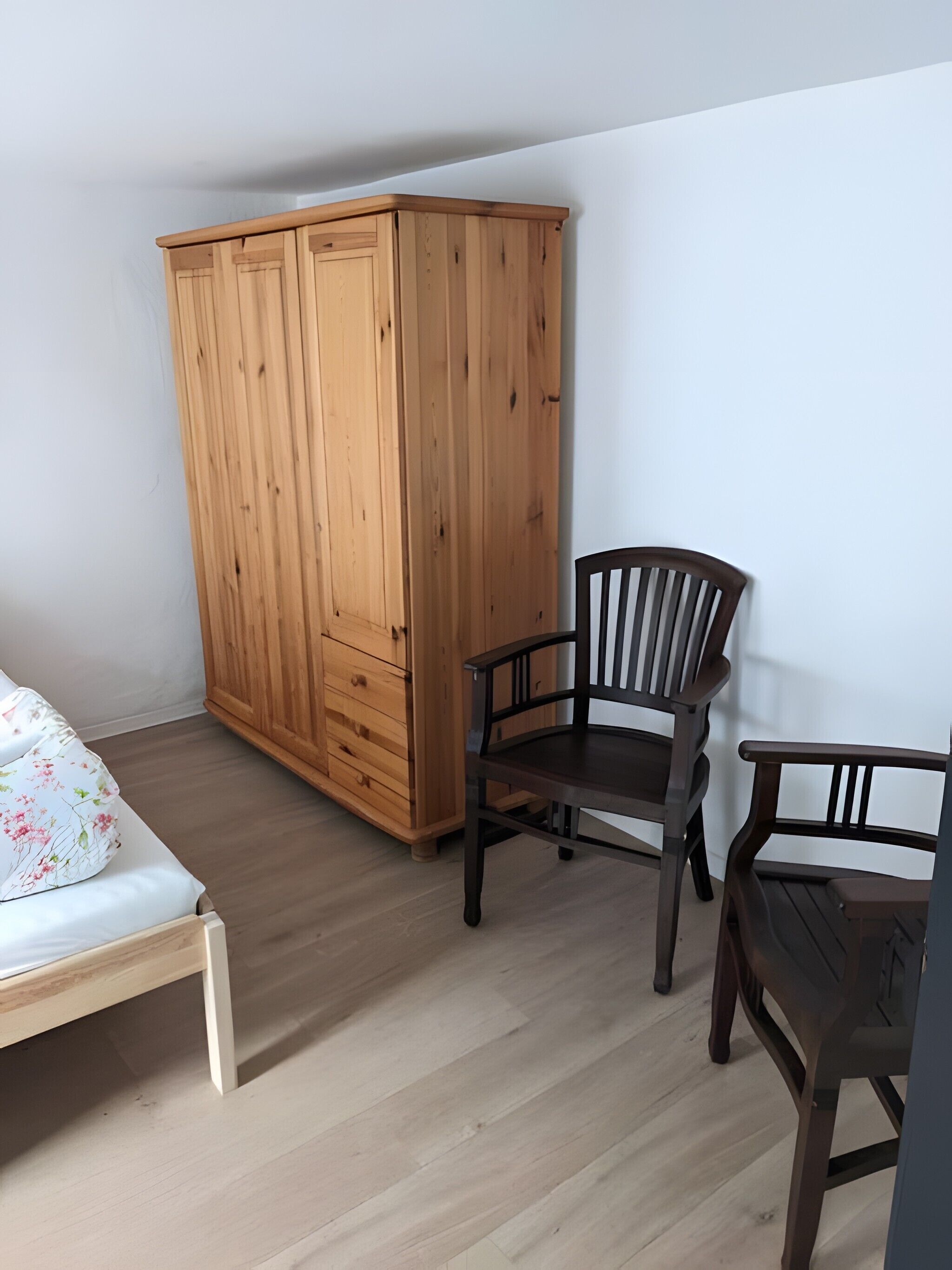 Ein Schlafzimmer mit einem Holzschrank, zwei Holzstühlen, einem Bett mit floralem Überwurf und einem hellen Holzboden.