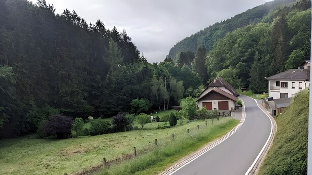 Una carretera rural serpentea por un paisaje verde con un edificio pequeño y una casa a la derecha.