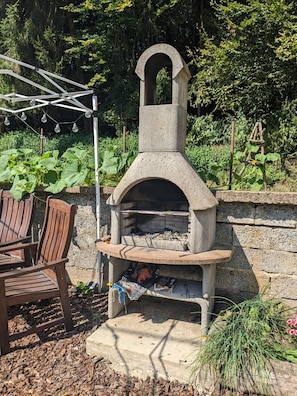 A stone outdoor oven with a chimney, surrounded by a garden with plants and a wooden chair. - Apartment for 4 People in the Eifel with Balcony and Garden (Baustert)