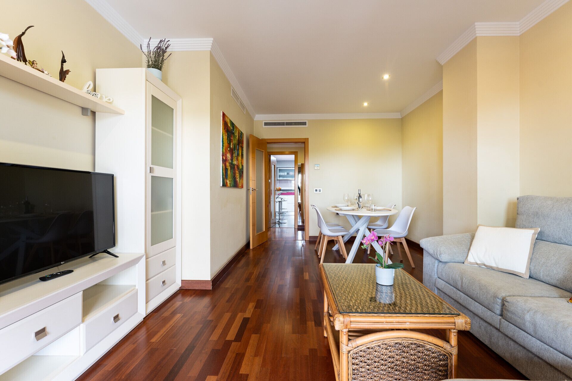 A living room with a television, sofa, dining table, chairs, and a hallway leading to a kitchen.