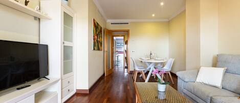 A living room with a television, sofa, dining table, chairs, and a hallway leading to a kitchen.
