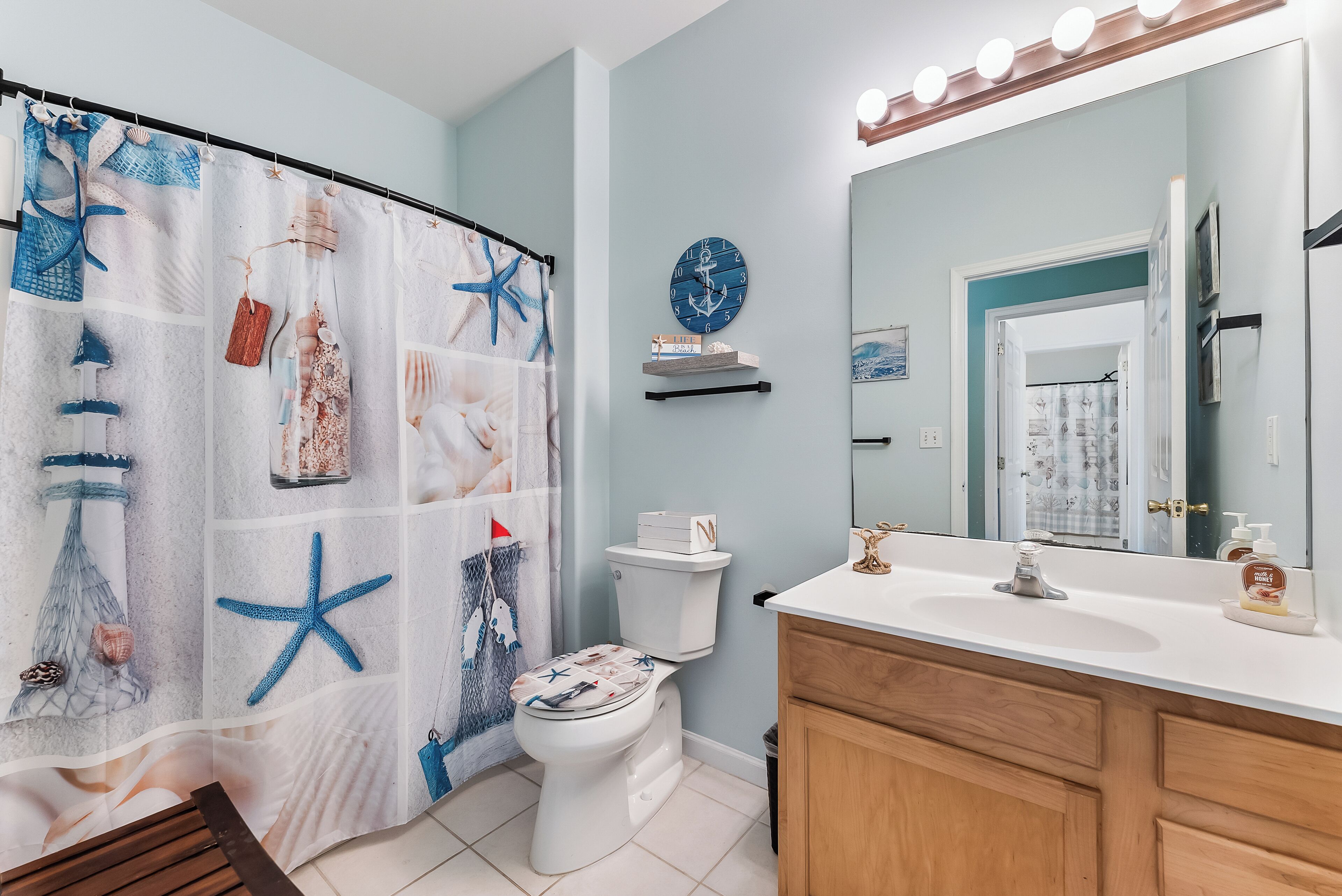 Une salle de bain avec un rideau de douche à motifs nautiques, des toilettes, un lavabo avec un miroir et une étagère avec des livres.
