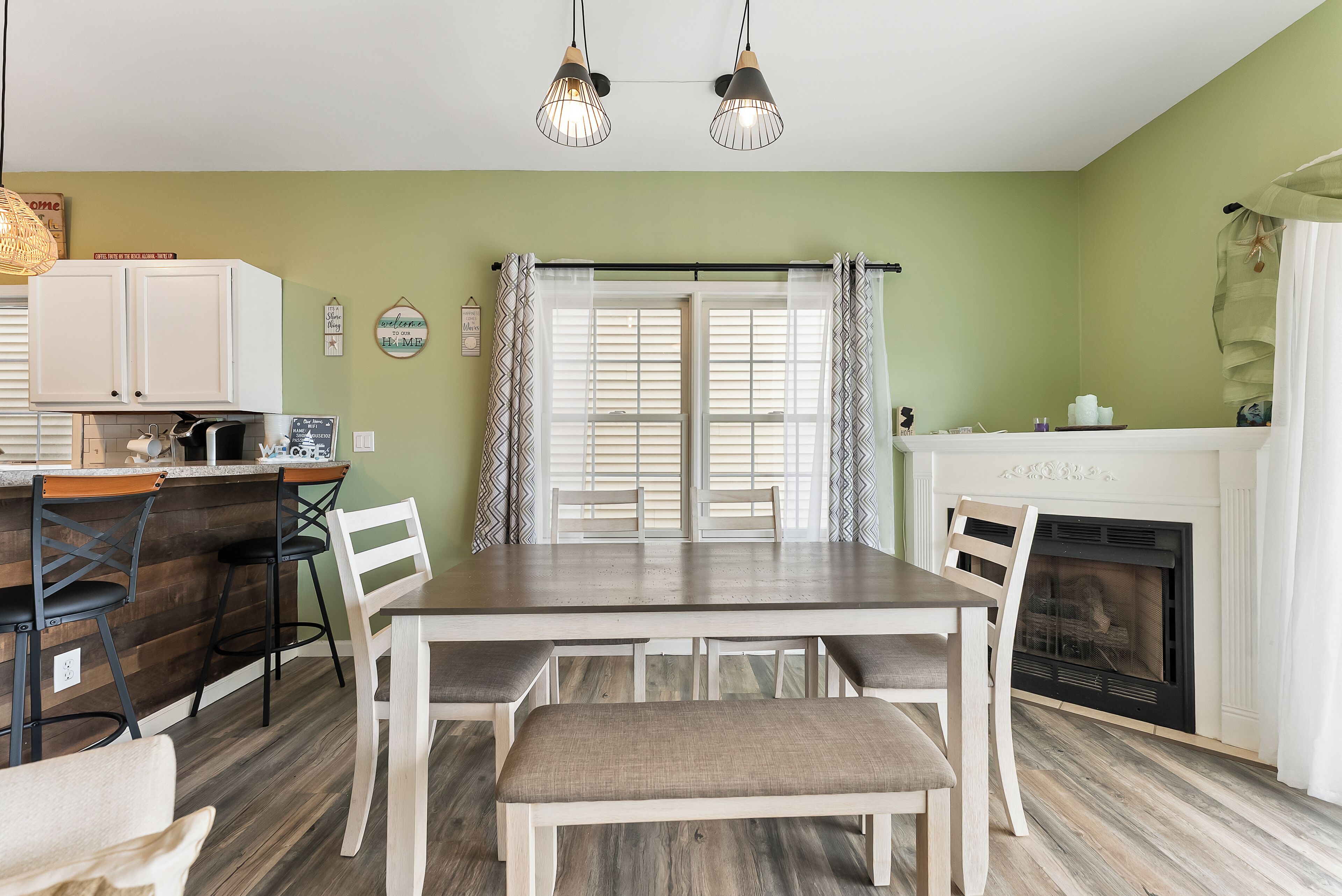 Un comedor con una mesa de madera, sillas blancas, un banco, una chimenea y una zona de cocina con mostrador y taburetes.
