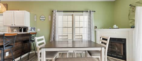 Un comedor con una mesa de madera, sillas blancas, un banco, una chimenea y una zona de cocina con mostrador y taburetes.