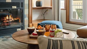 Ein gemütliches Wohnzimmer mit Kamin, einem runden Holztisch mit Snacks, einem blauen Kissen und Blick ins Freie.