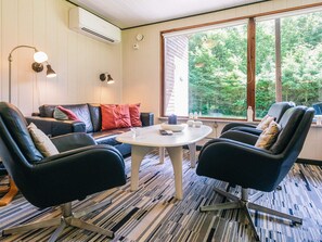 Ein Zimmer mit einer Couch, zwei Sesseln, einem Tisch und einem großen Fenster mit Blick auf Bäume.