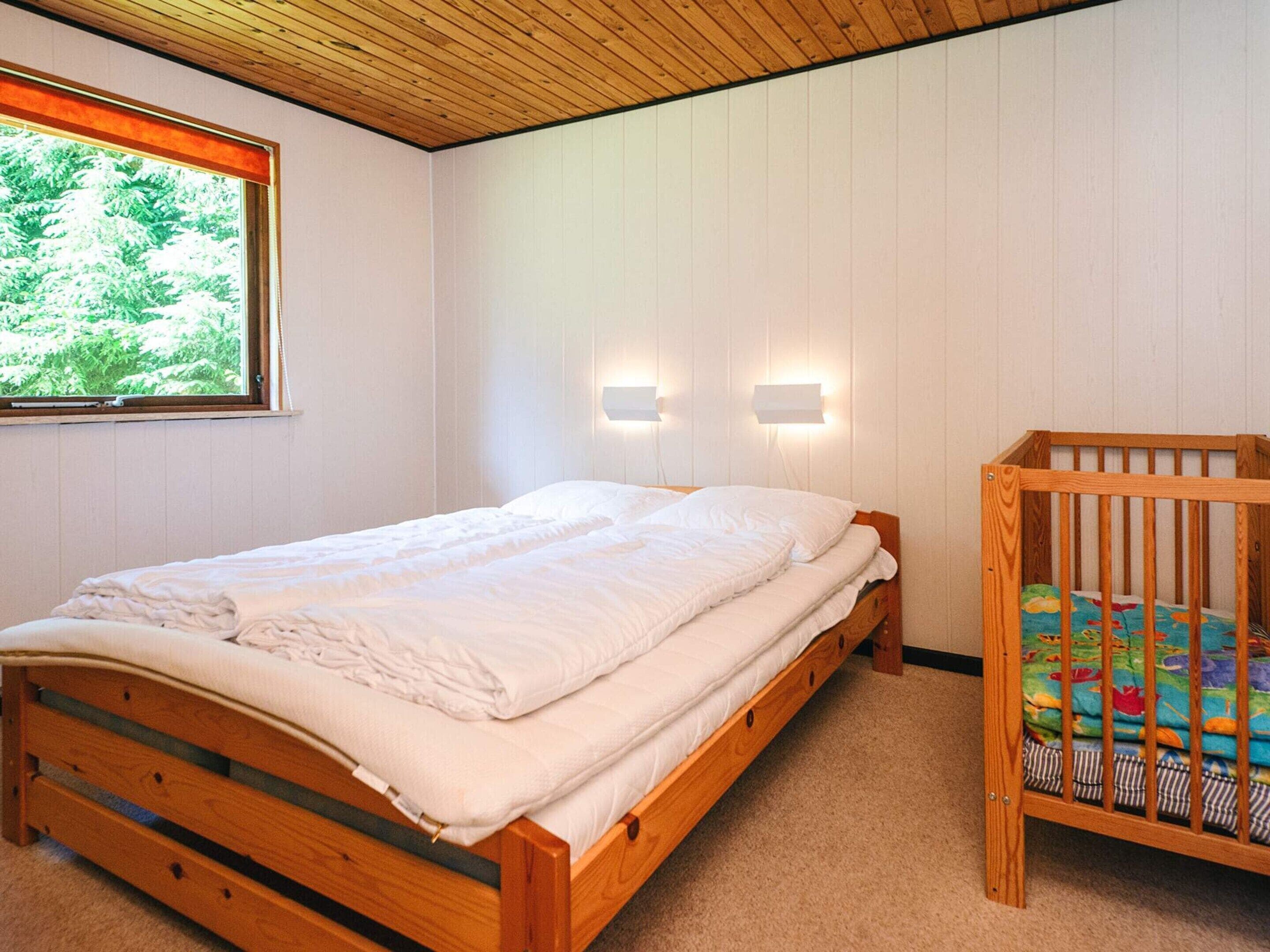 A bedroom with a wooden bed, a crib, a window with a view of greenery, and wall-mounted lights.