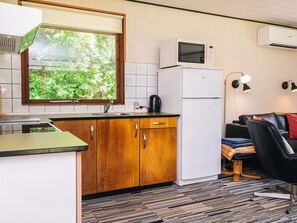 Eine kompakte Küche mit Mikrowelle, Kühlschrank und Spüle. Ein Fenster bietet Blick auf Grünflächen. Im Wohnbereich befinden sich eine Couch und eine Lampe.