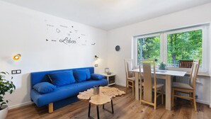Habitación con sofá azul, mesa de comedor de madera, sillas, una mesita auxiliar, una planta en maceta y ventana con vista a árboles.