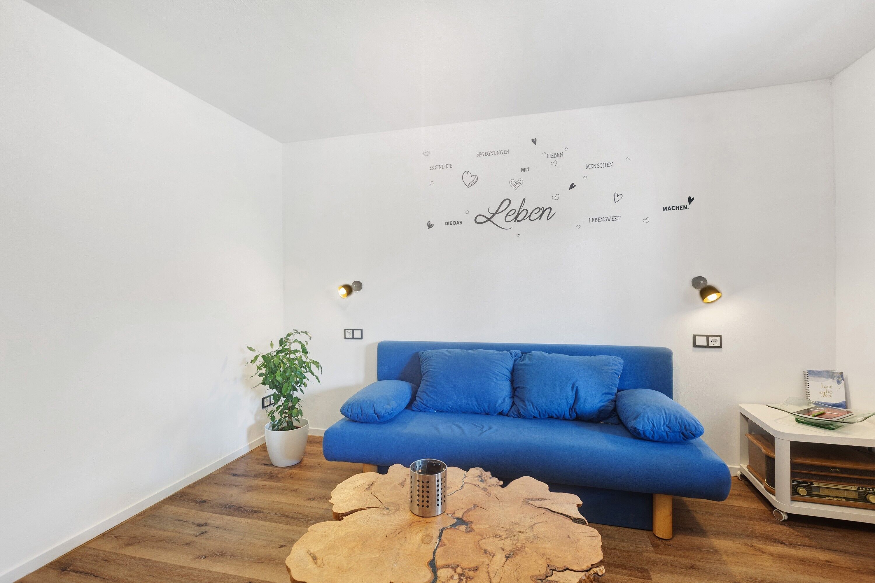 Een moderne woonkamer met een blauwe bank, een houten salontafel, een potplant en een muur met motiverende tekst.