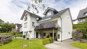Una casa moderna de dos pisos con porche cubierto, paneles solares y entrada pavimentada.