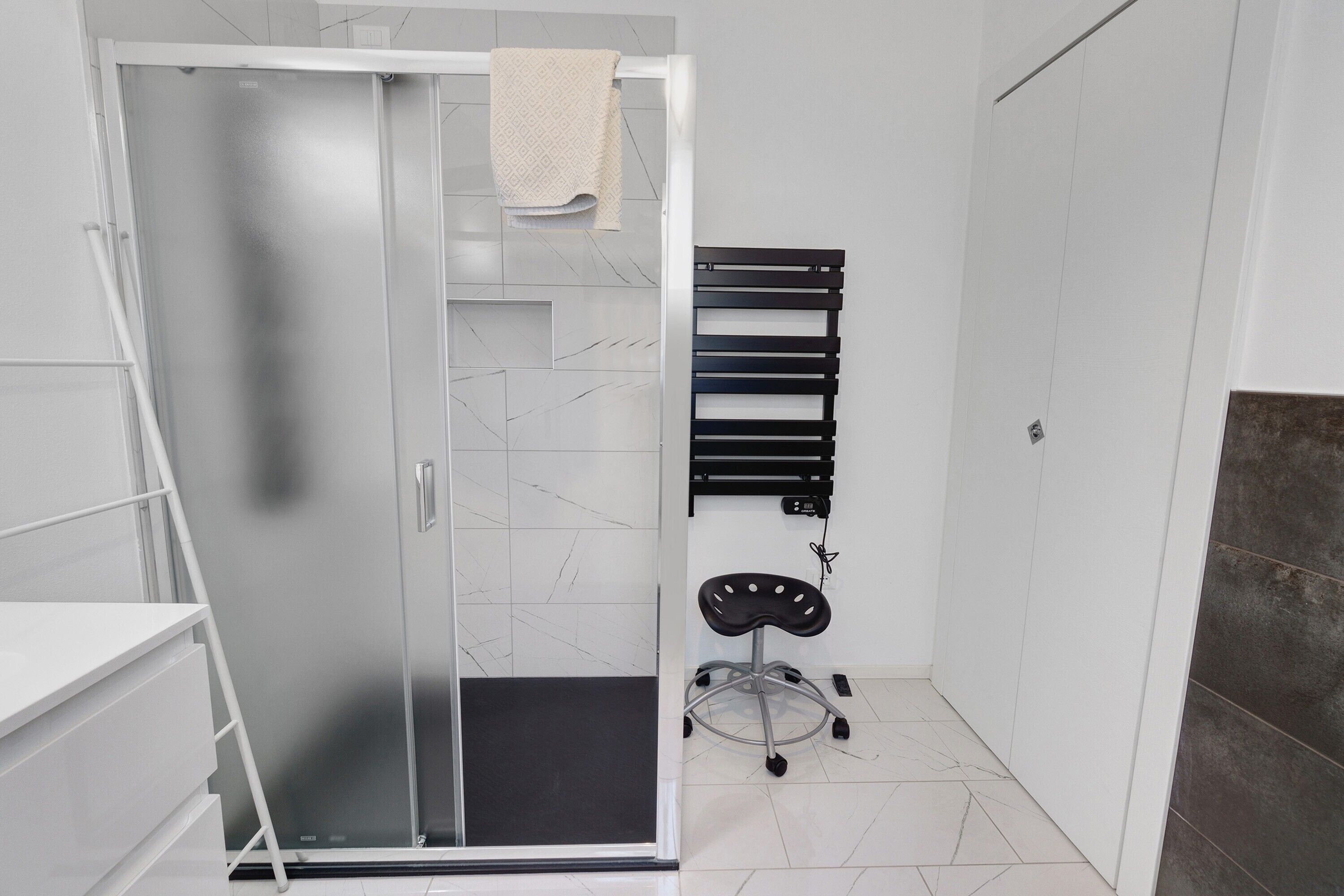 Un baño con cabina de ducha de vidrio, un perchero, una silla negra y suelo de baldosas.