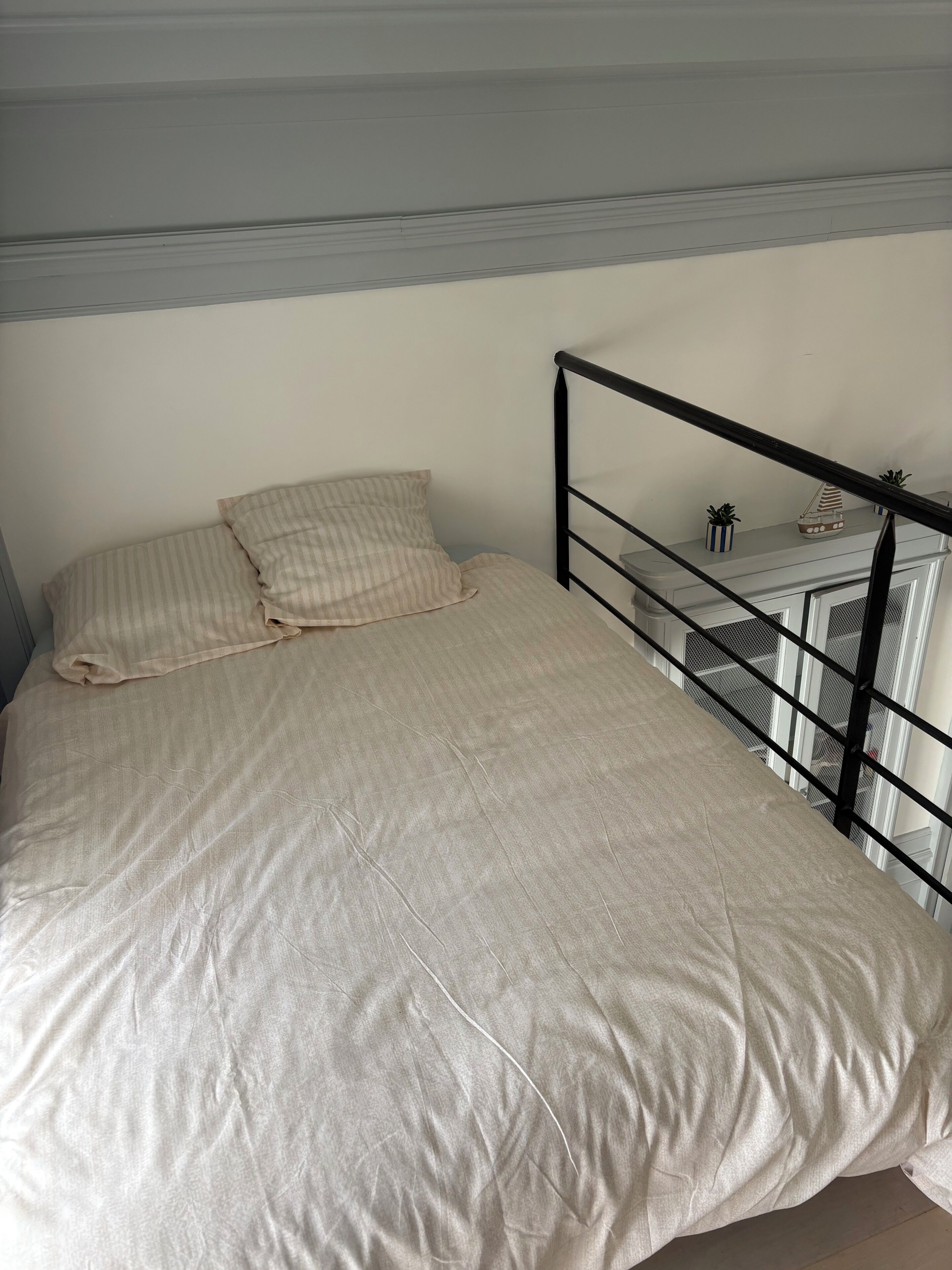 Un lit avec un oreiller à rayures, une tête de lit en métal et une petite étagère ornée de plantes en pot et d’un modèle de bateau.