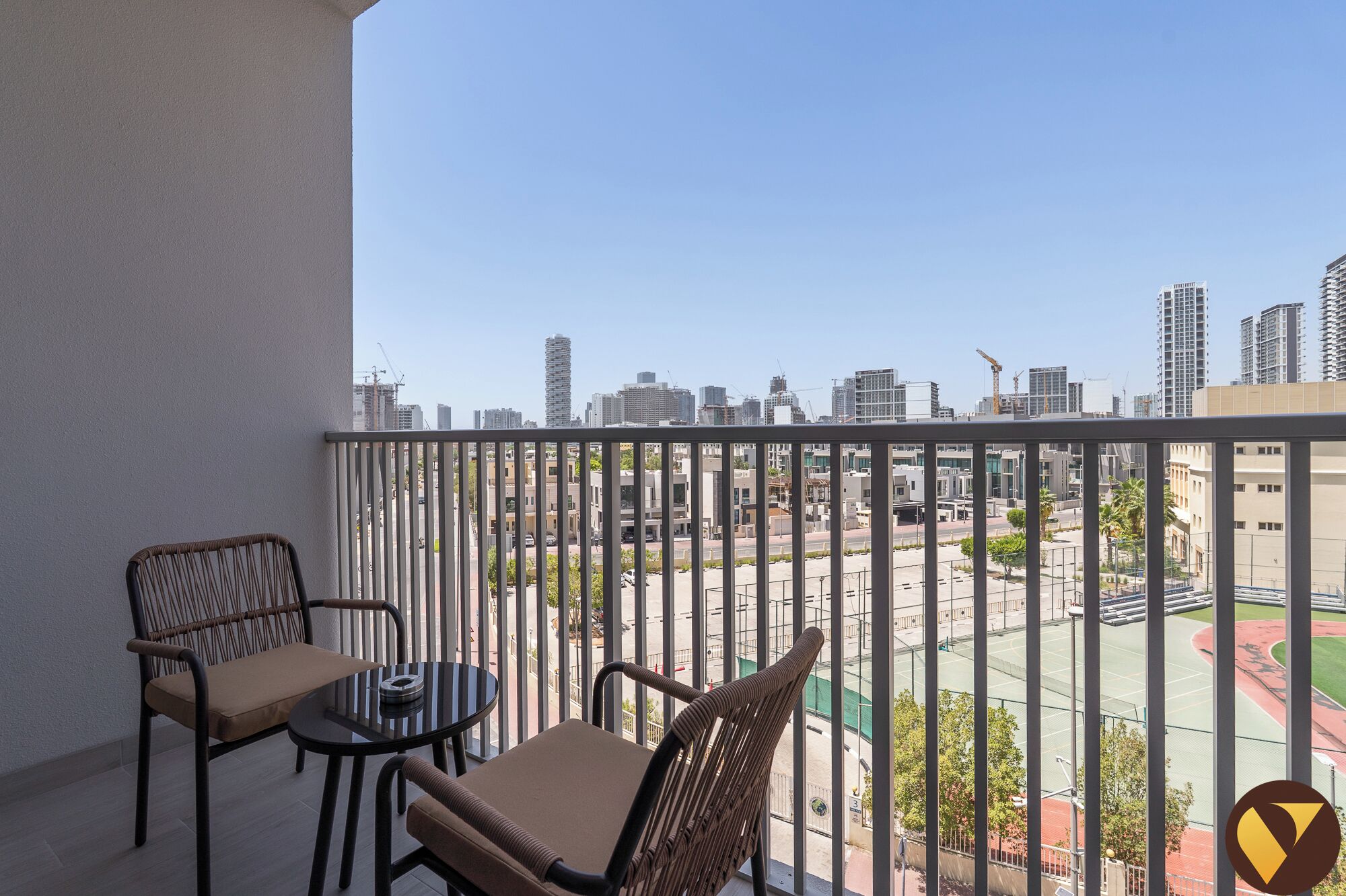 A balcony with two wicker chairs and a small table, overlooking a cityscape with buildings and a sports court.