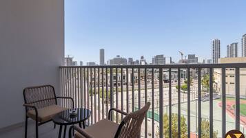 A balcony with two wicker chairs and a small table, overlooking a cityscape with buildings and a sports court.