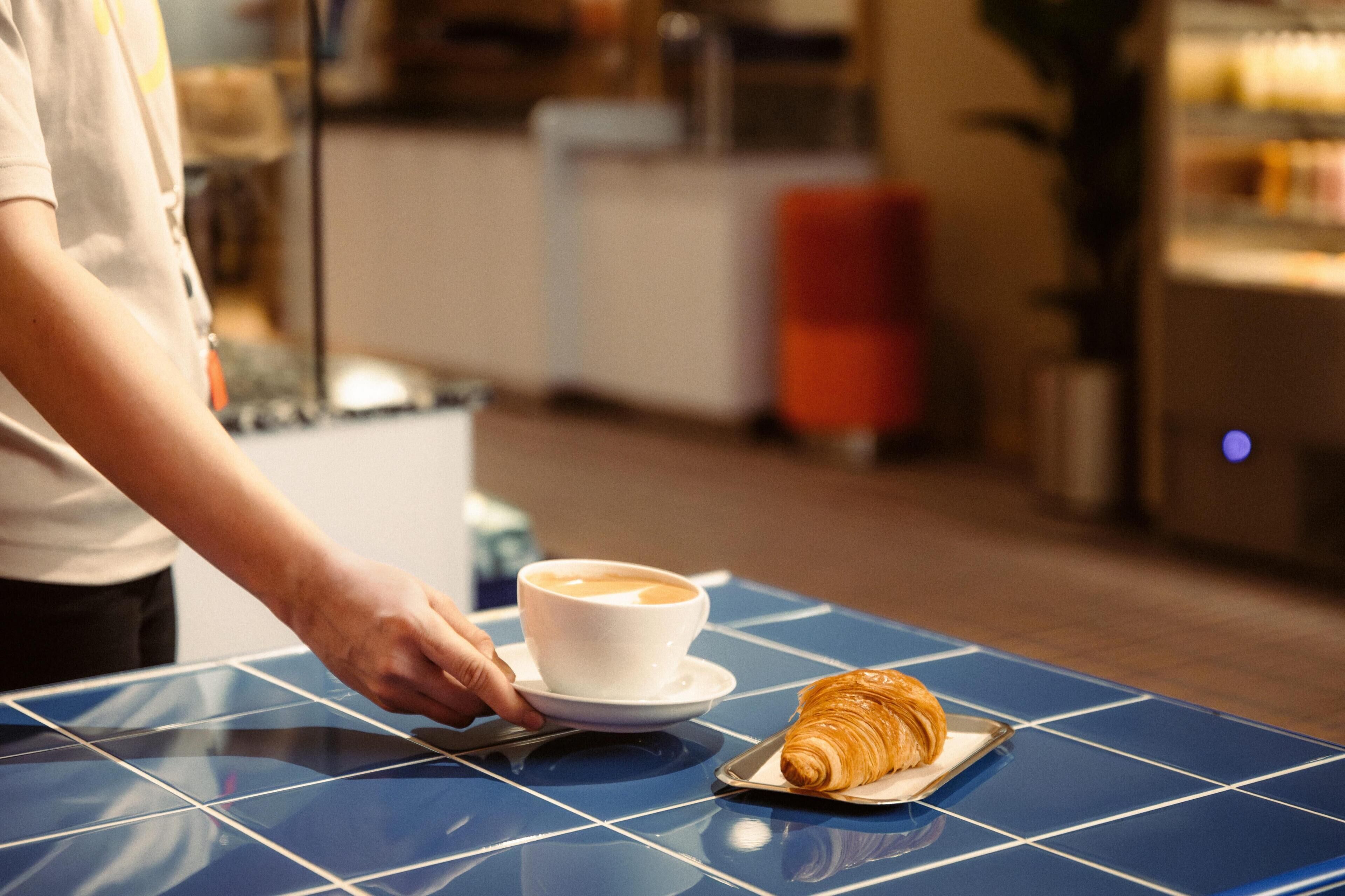 A person serving a cup of coffee and a croissant on a blue-tiled table.