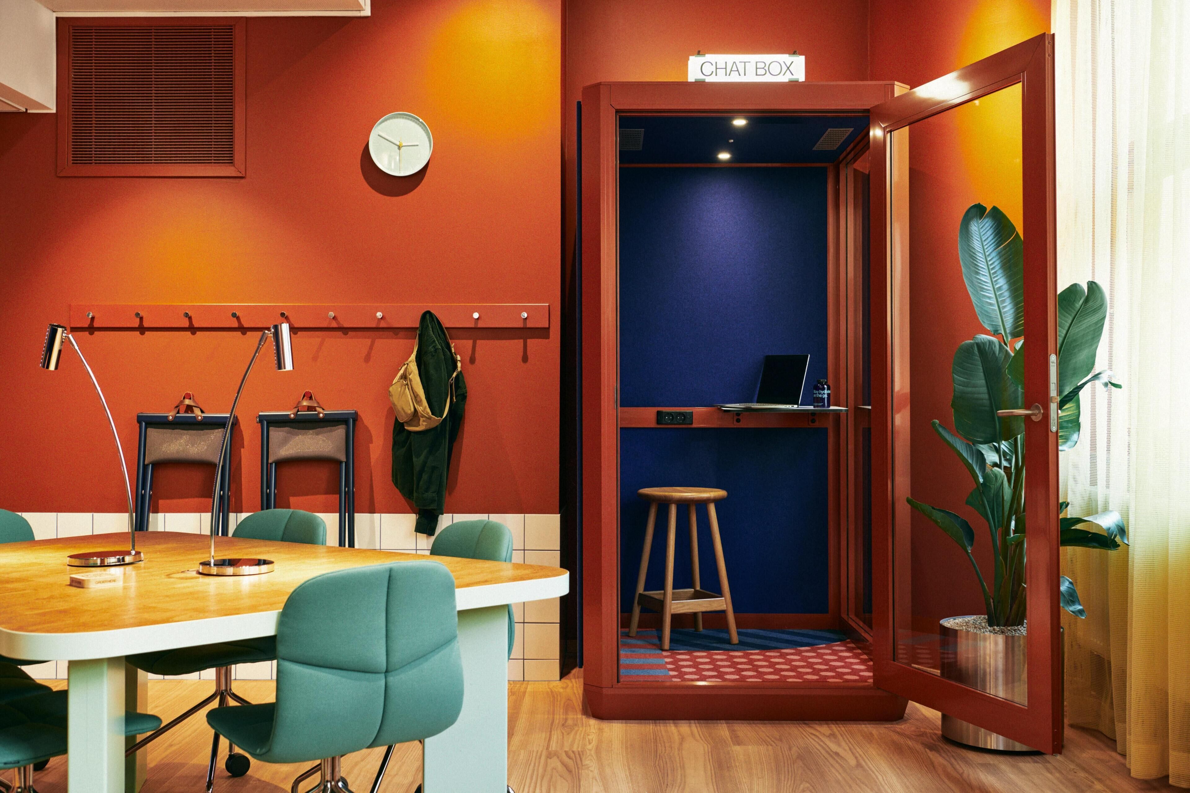 A conference room with a wooden table, four chairs, a clock, a coat rack, and a chat box.