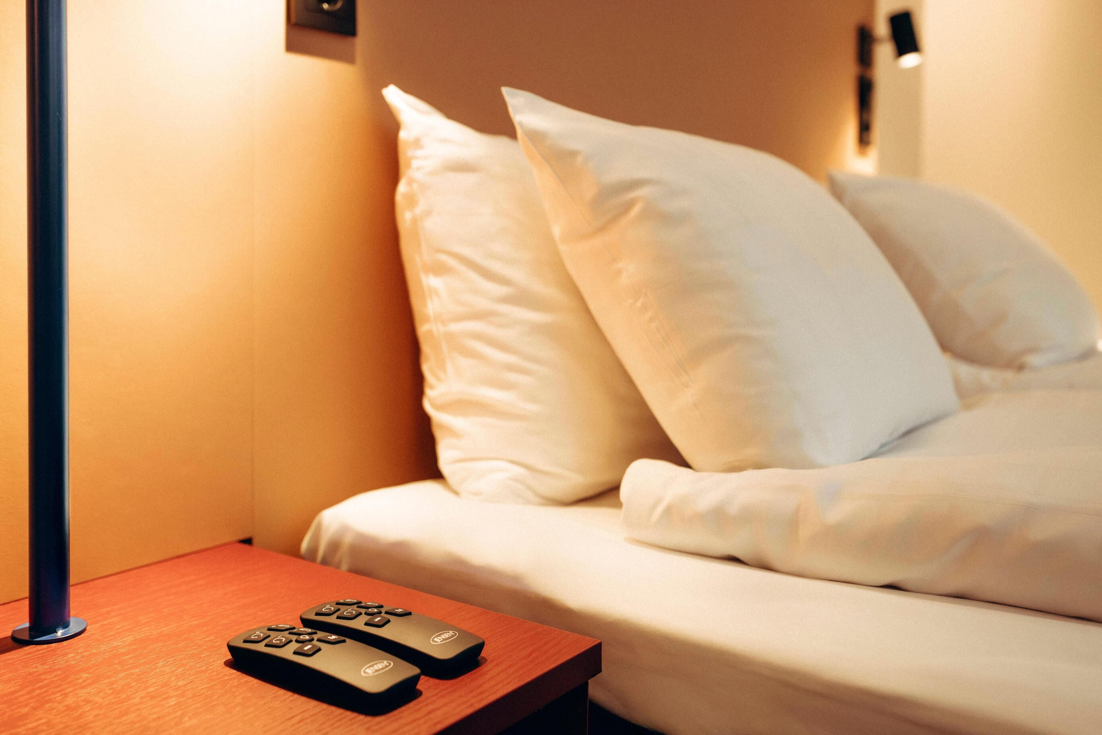 A hotel room with a bed, pillows, a nightstand with a remote control, and a wall-mounted lamp.