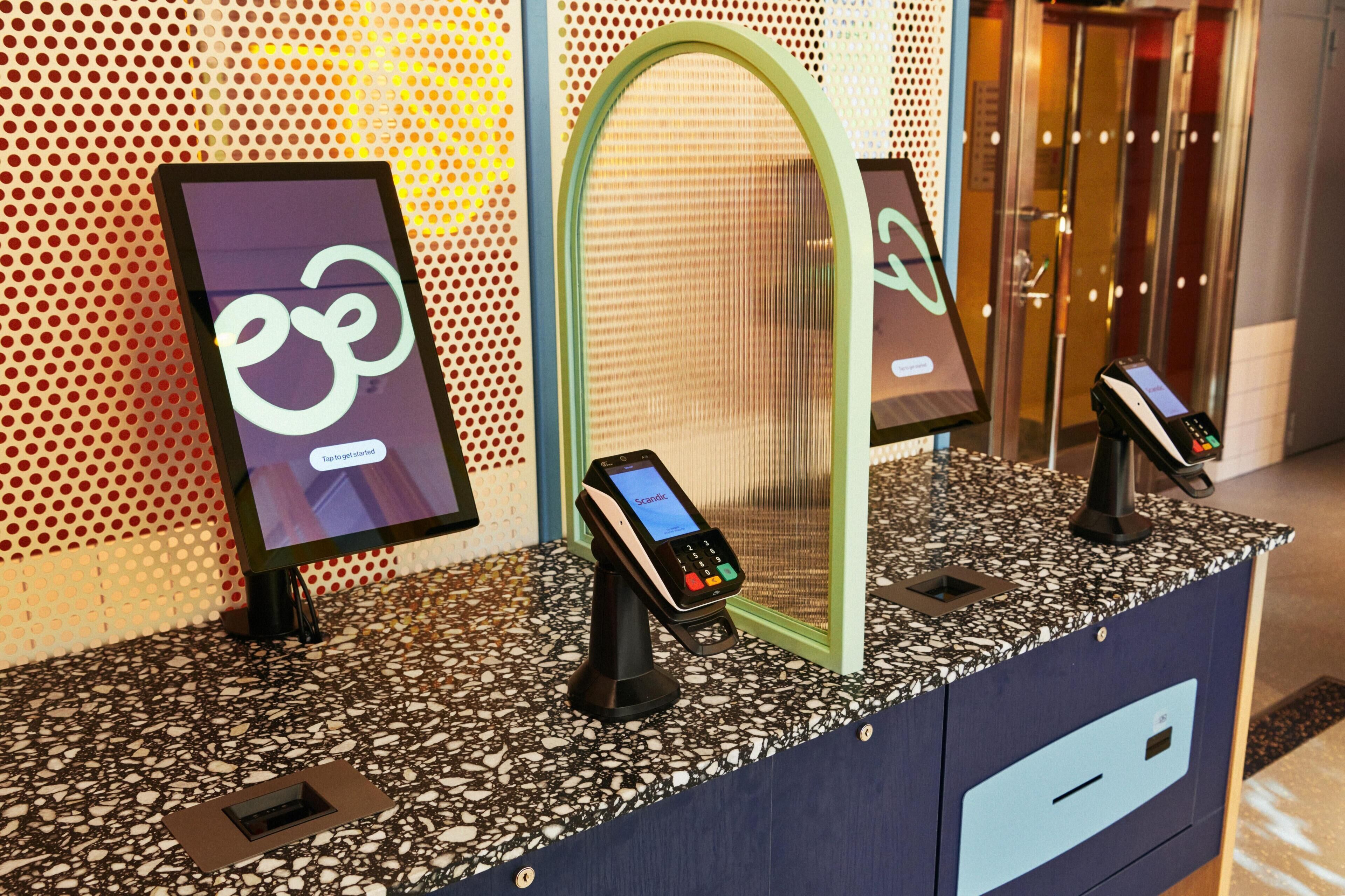 Quiosco de autoservicio con una pantalla que muestra un logotipo y un mensaje, un terminal de pago y una estructura con espejo.