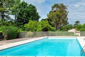 A rectangular swimming pool with a white fence and metal ladder, surrounded by lush green trees and a clear sky.