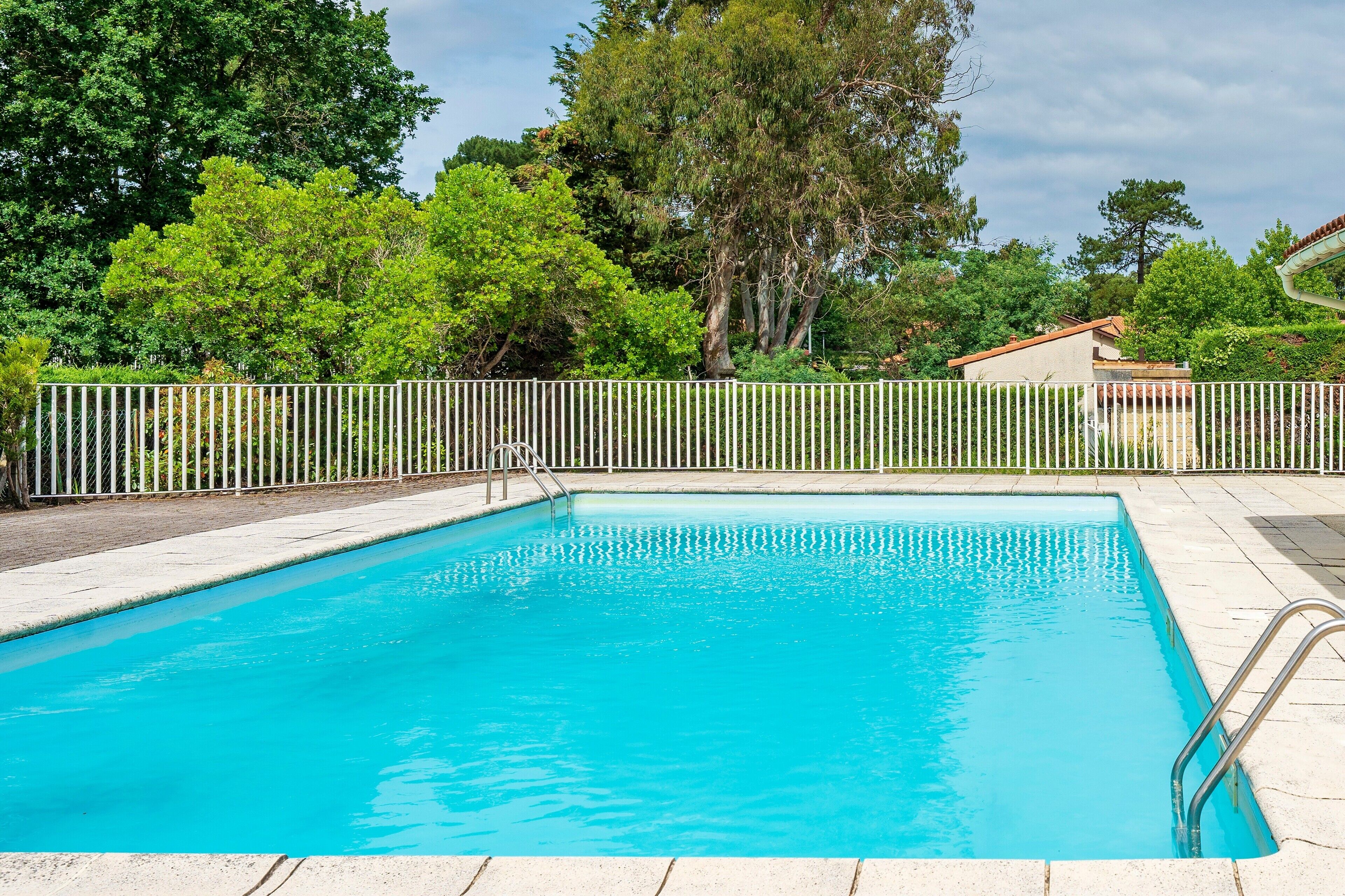 A rectangular swimming pool with a metal ladder on one side, surrounded by a white fence, with trees and a building in the background.