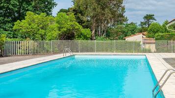 A rectangular swimming pool with a metal ladder on one side, surrounded by a white fence, with trees and a building in the background.
