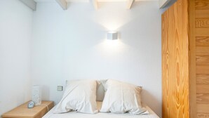 Een slaapkamer met een houten nachtkastje, een witte bijzettafel, een bed met witte lakens en een wandlamp.