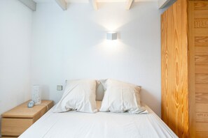 A bedroom with a wooden nightstand, a white bedside table, a bed with white bedding, and a wall-mounted light fixture.