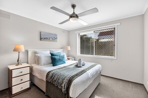 Une chambre à coucher comprenant un lit, des tables de chevet, un ventilateur de plafond, une fenêtre avec des stores et une œuvre d’art accrochée au mur.