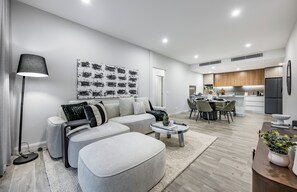 A modern living room with a sofa, coffee table, and lamp. A dining area with chairs and a table is visible in the background. - The Signature: 3BR, Crown & Stadium, Rooftop BBQ (Burswood)
