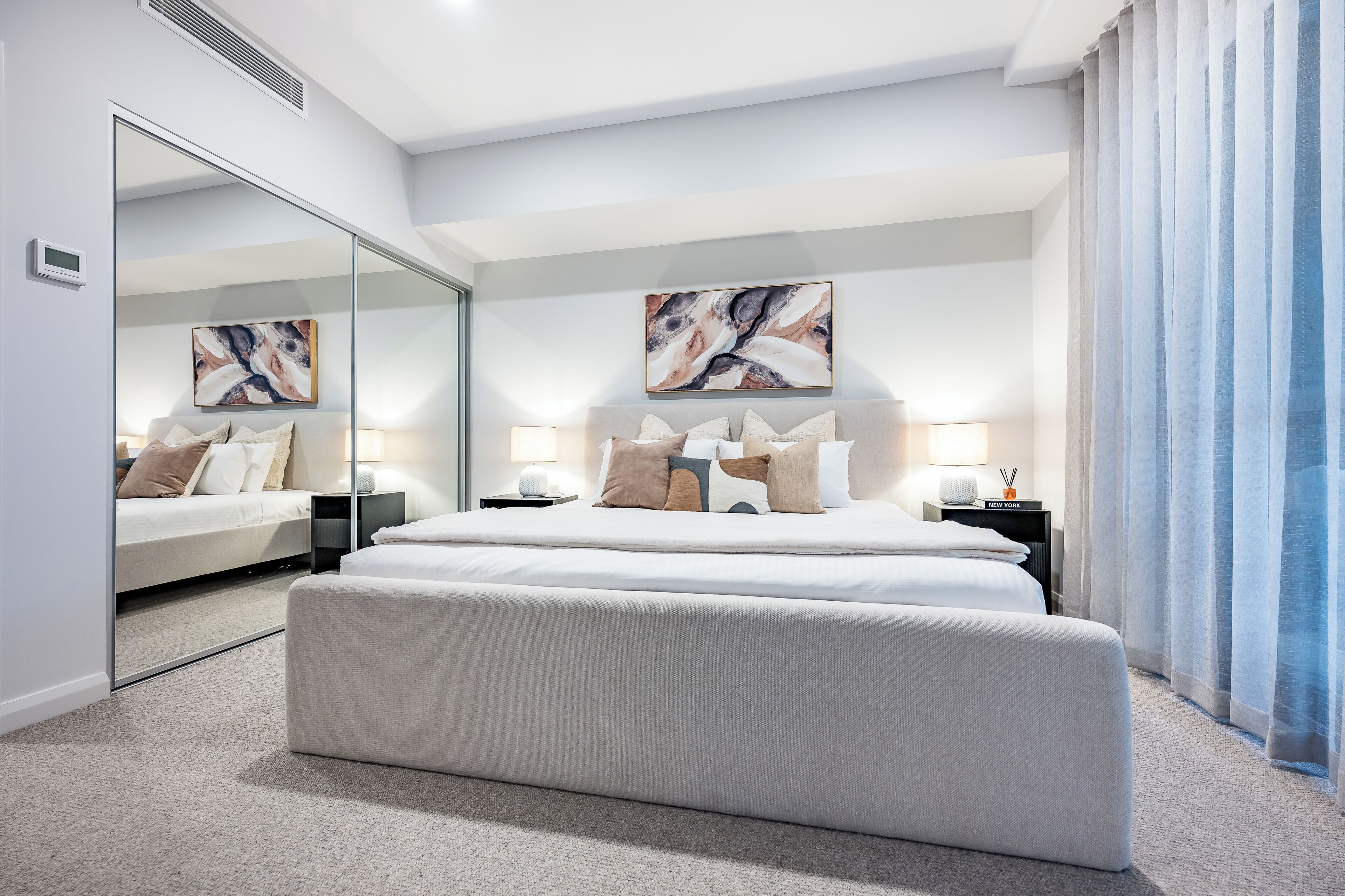 A modern hotel room with a large bed, bedside tables, a mirror, and abstract artwork on the wall.