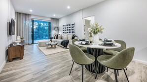 Una sala de estar moderna con sofá, mesa de comedor y sillas, televisor montado en la pared y un ventanal con vistas a árboles.