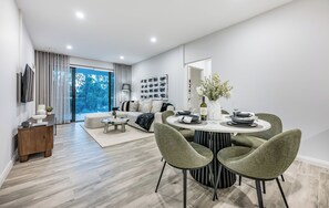A modern living room with a sofa, dining table, and chairs, a TV mounted on the wall, and a large window with a view of trees. - Luxury Burswood Stay · Crown, Stadium & Swan River · Sleeps 6 (Burswood)