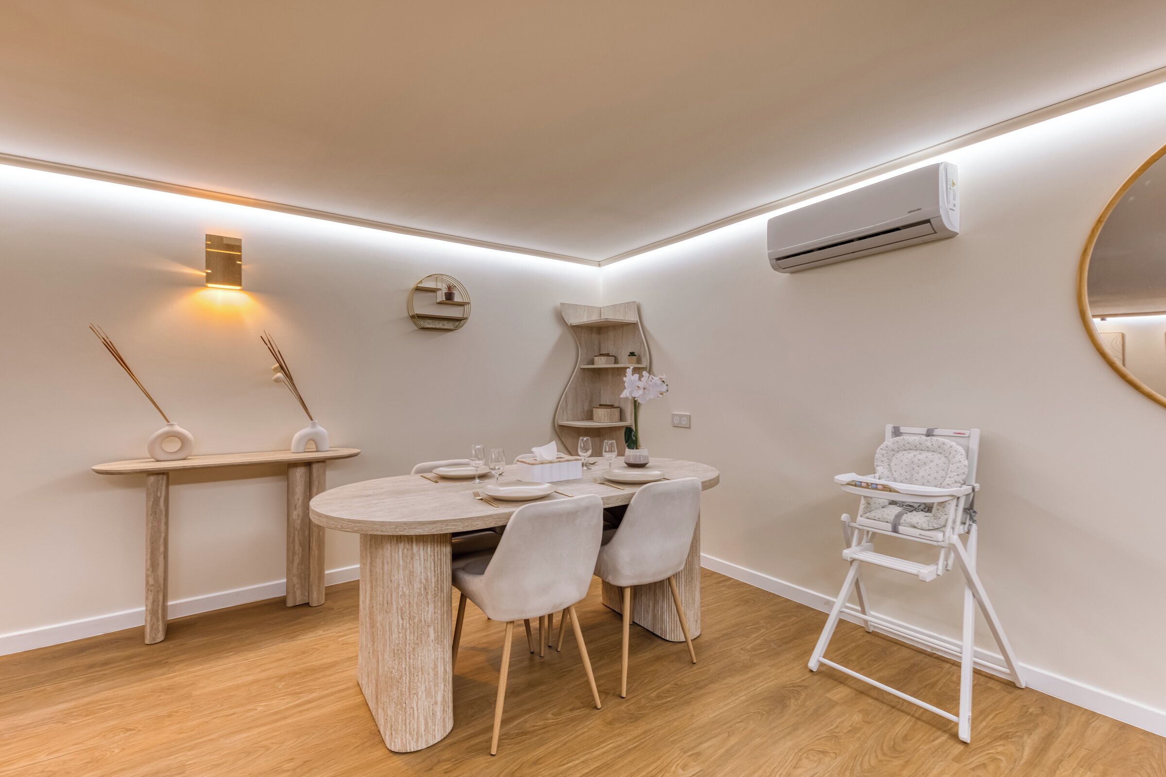 Een eetruimte met een ronde tafel, stoelen, een kinderstoel, een houten bijzettafeltje, wandplanken en een airconditioner.