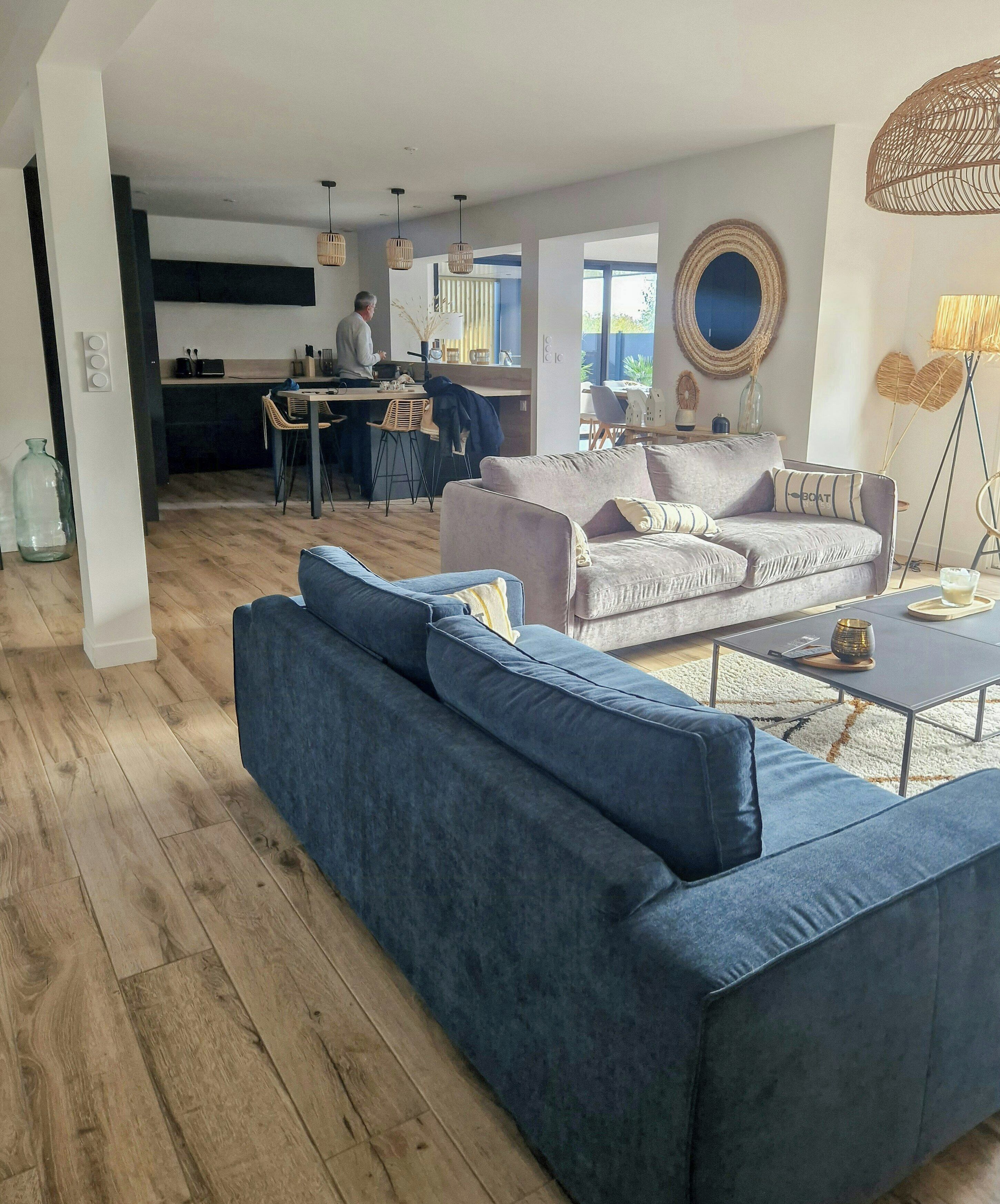 Ein modernes Wohnzimmer mit einem blauen Sofa, einem grauen Sofa und einem Couchtisch. Im Hintergrund ist eine Küchenzeile erkennbar, wo jemand kocht.