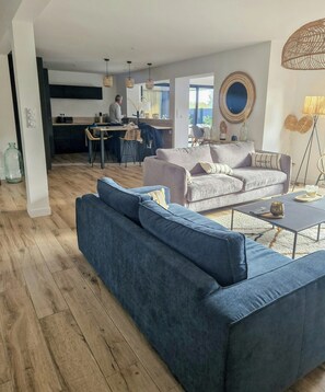 Ein modernes Wohnzimmer mit einem blauen Sofa, einem grauen Sofa und einem Couchtisch. Im Hintergrund ist eine Küchenzeile erkennbar, wo jemand kocht.