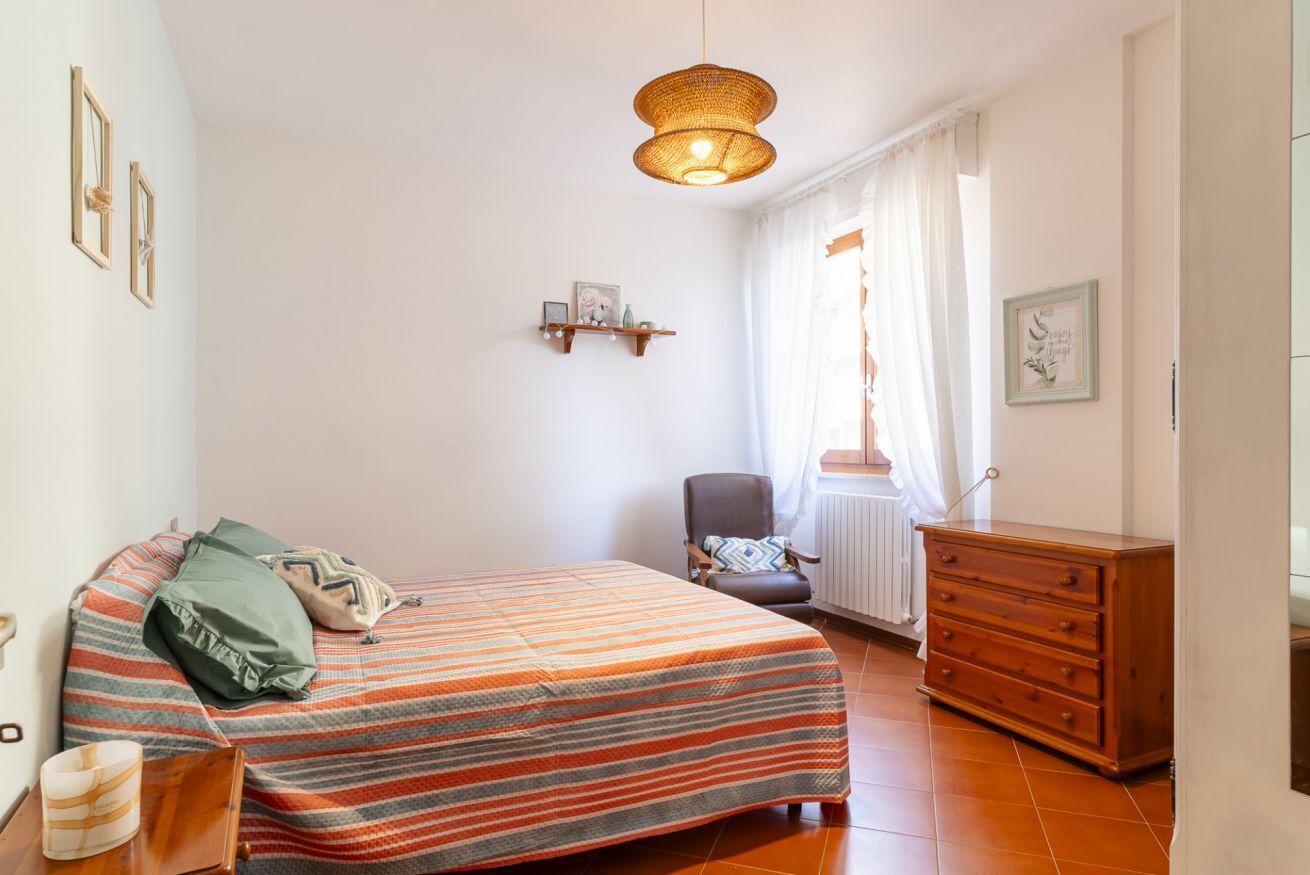 Een slaapkamer met een bed, een stoel, een dressoir, een plank met spullen en een hanglamp.