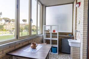 Una stanza con una finestra, un tavolo con una tazza, uno scaffale bianco con oggetti da cucina, un lavandino e un piccolo armadietto.