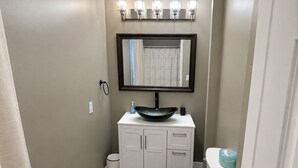 A bathroom with a white toilet, a white vanity with a black sink, a mirror above the vanity, and a light fixture above the mirror. - 2-bedroom apartment in Lewiston near CMCC (Lewiston)