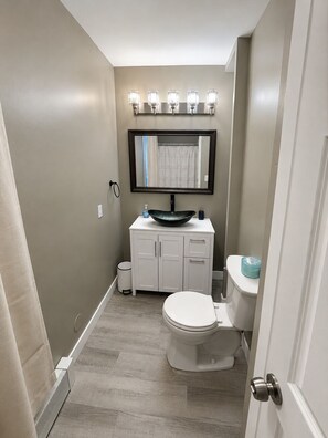 A bathroom with a white toilet, a white vanity with a black sink, a mirror above the vanity, and a light fixture above the mirror. - 2-bedroom apartment in Lewiston near CMCC (Lewiston)