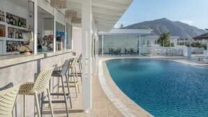 Zona de bar junto a la piscina, con sillas, mostrador de bar con botellas y vista a la montaña.
