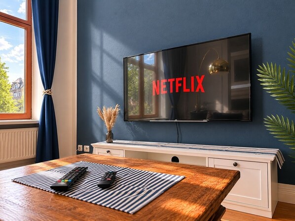 Ein Zimmer mit einem großen Flachbildfernseher an der Wand, einem Holztisch mit Fernbedienungen, einer Vase mit Trockenpflanzen und einem Fenster mit blauen Vorhängen.