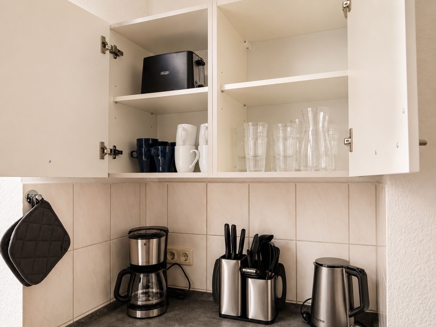 Eine Küche mit Kaffeemaschine, Wasserkocher, einem Messerblock sowie einem Schrank mit Tassen und Gläsern.