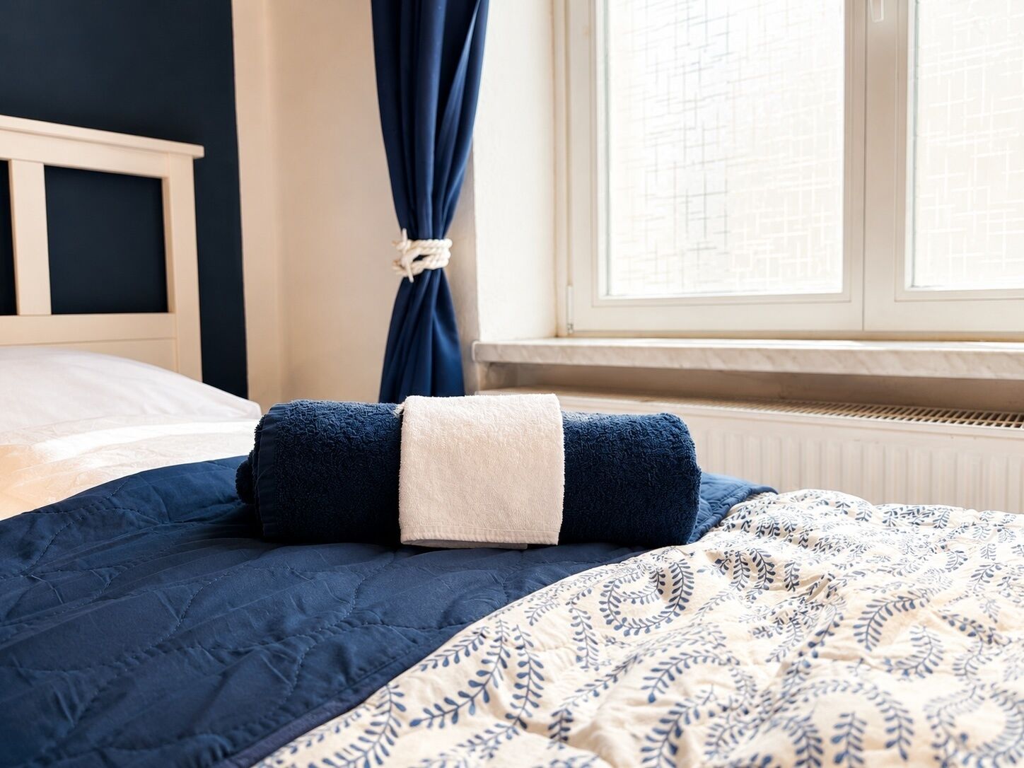 Un letto con una coperta azzurra, una trapunta bianca e una tenda a motivi.